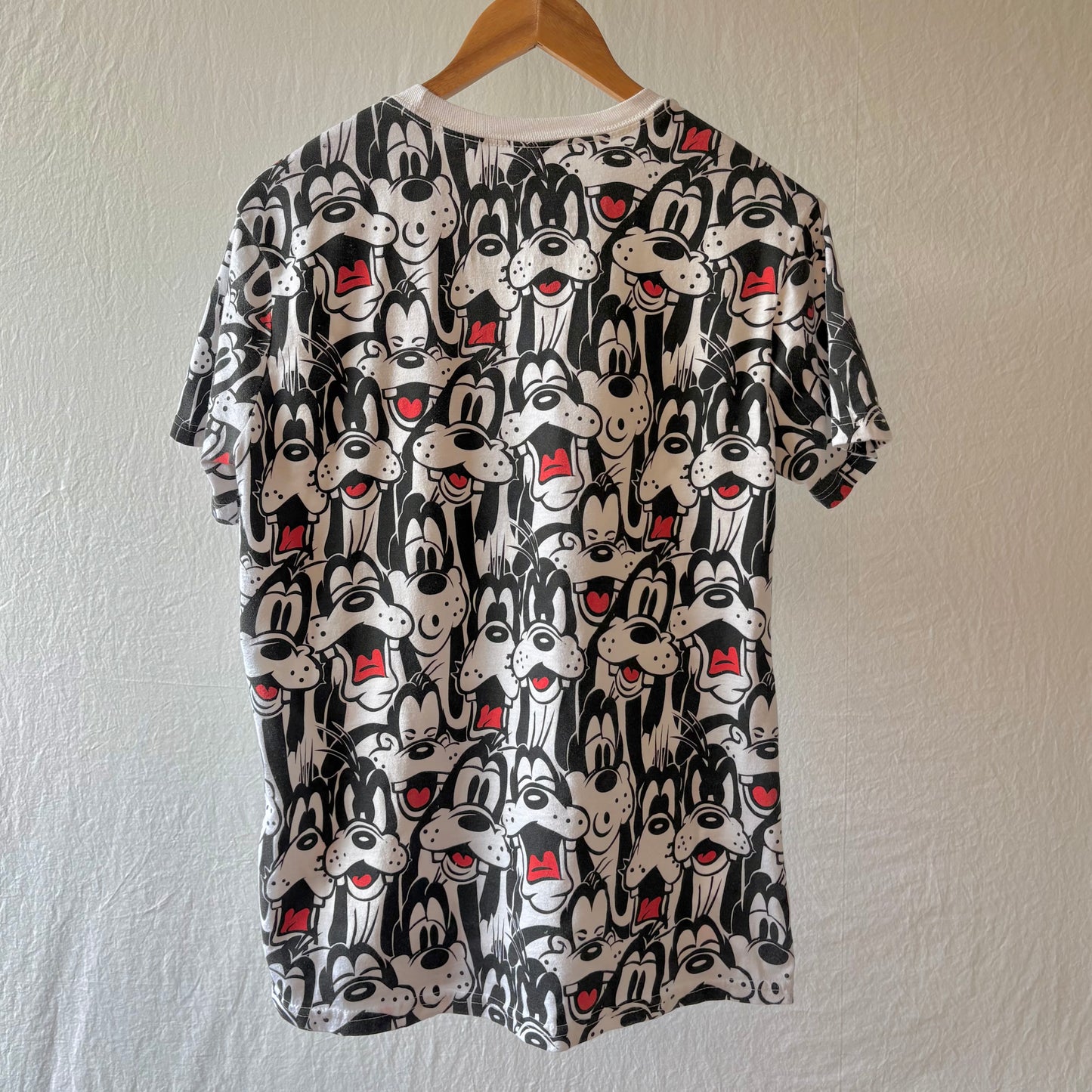A short-sleeve crewneck t-shirt hanging on a wooden hanger against a textured off-white wall. The shirt features an all-over repeating print of Goofy faces in black and white with small red accents.