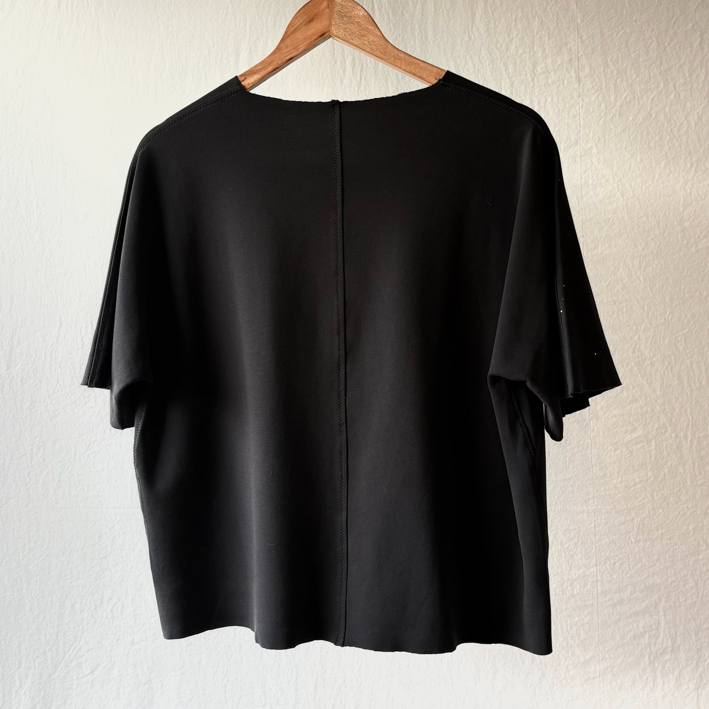 A black short-sleeve t-shirt hanging on a wooden hanger against a textured off-white wall, showing the back view with a central seam.