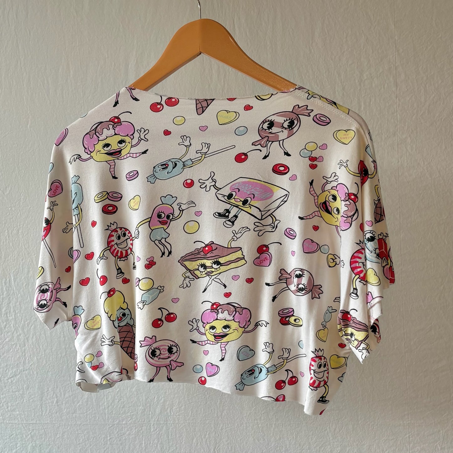 A short-sleeve crop top with a white base and a colorful cartoon print of desserts, characters, and hearts, hanging on a wooden hanger against a textured off-white wall.