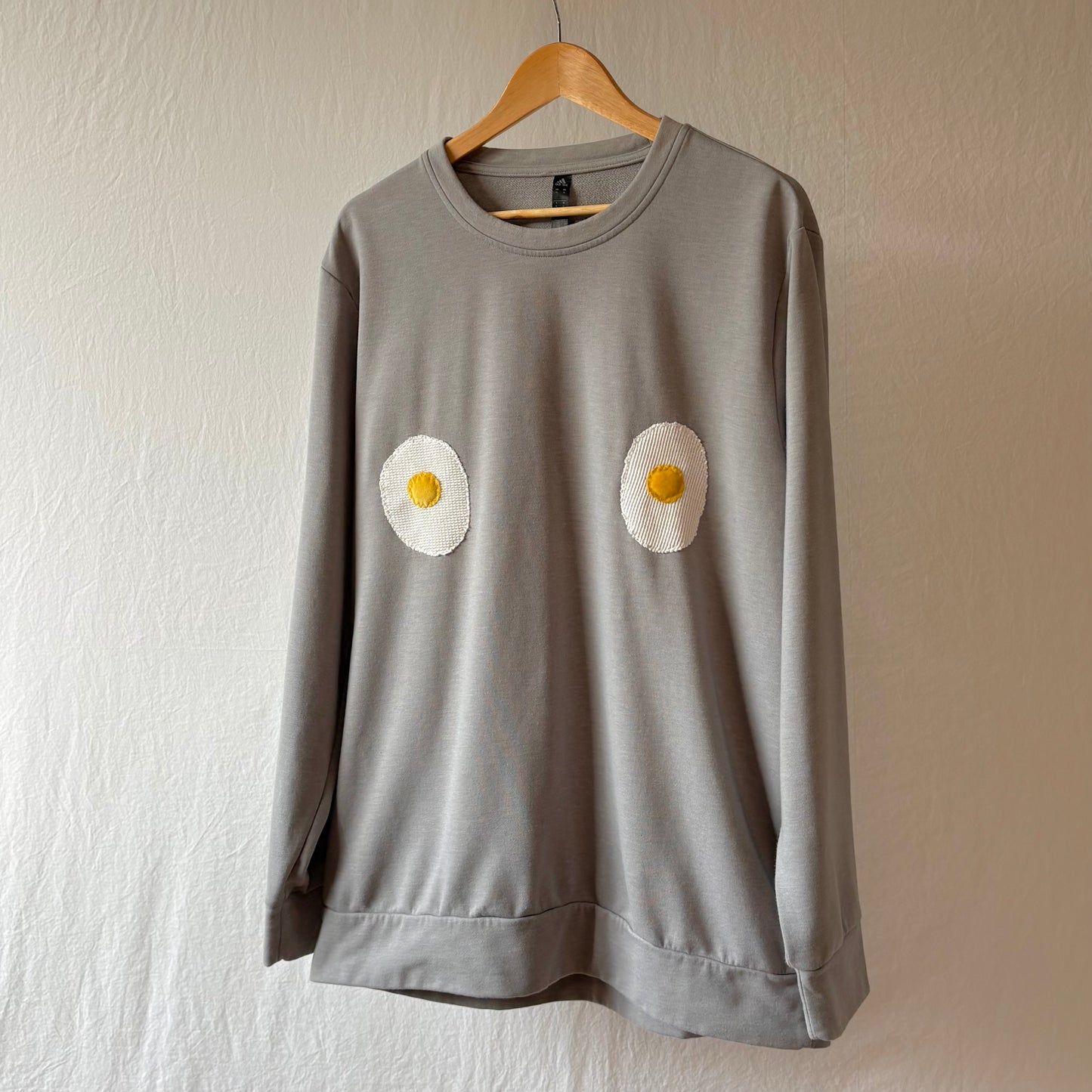 A light grey long-sleeve sweatshirt hanging on a wooden hanger against a textured off-white wall. The sweatshirt features two handmade egg patches on the chest.