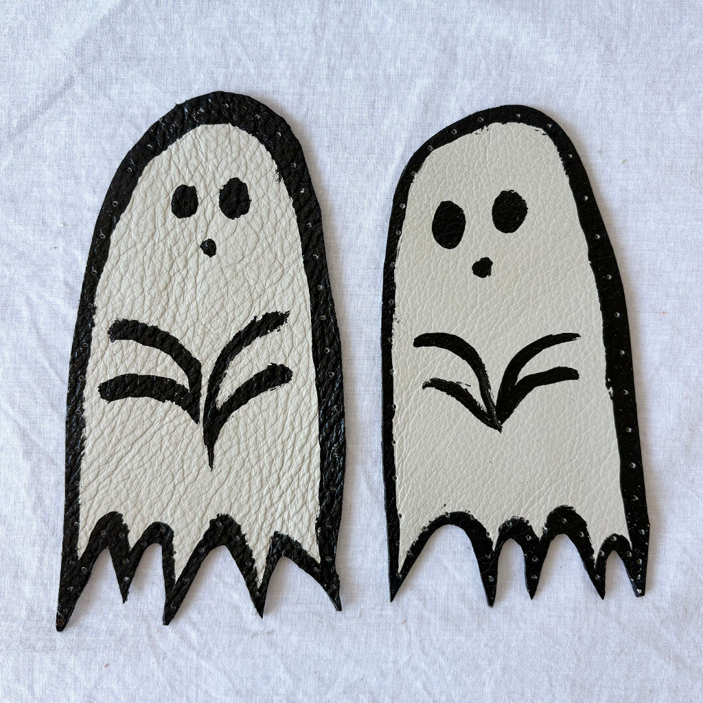 Two white ghost-shaped white leather patches with black outlines, with holes punched around the edges.  on a white background