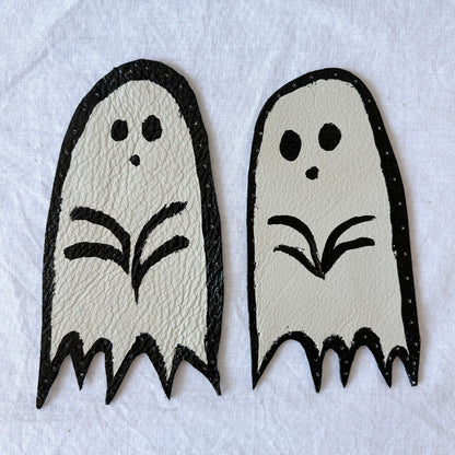 Two white ghost-shaped white leather patches with black outlines, with holes punched around the edges.  on a white background