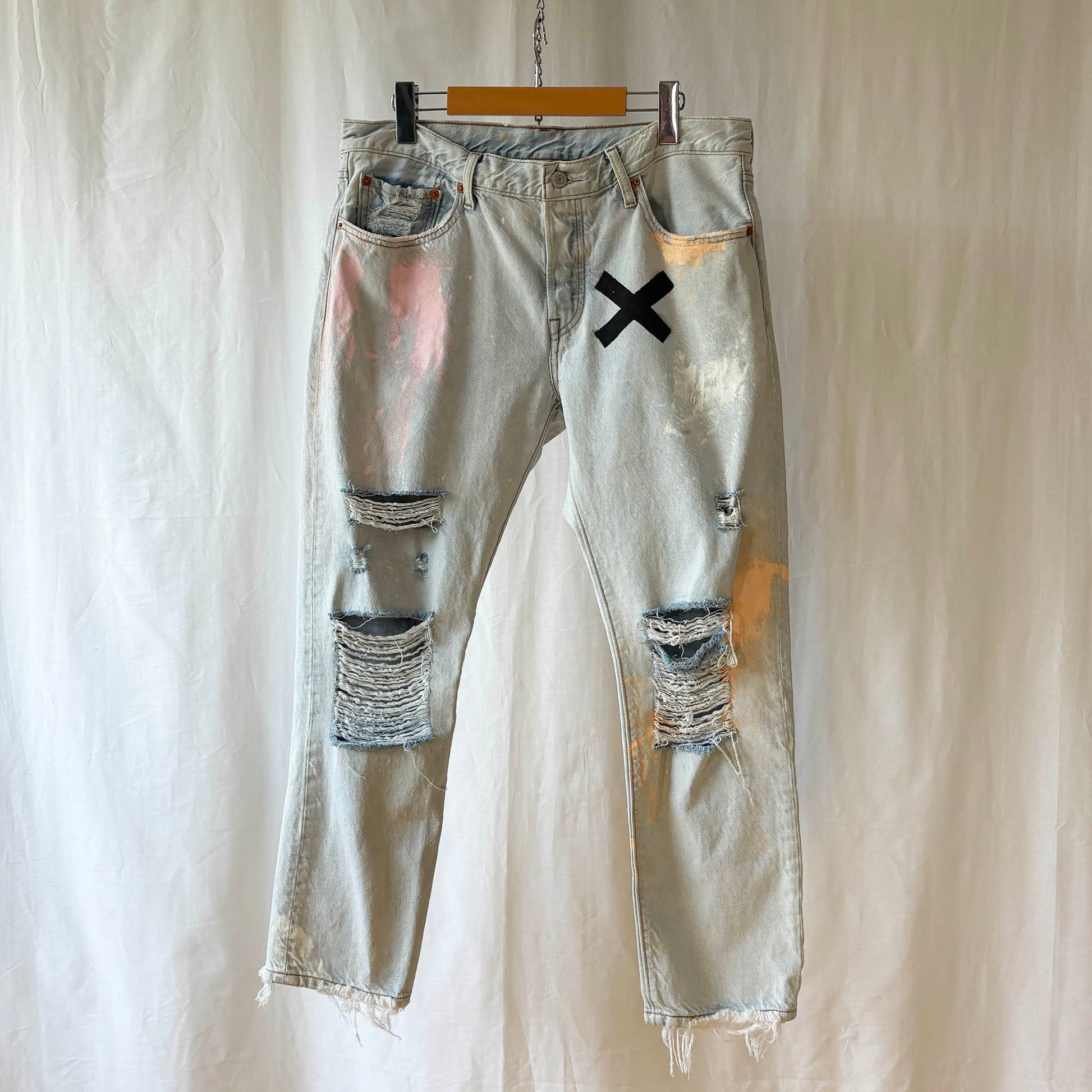 Ripped light blue jeans with a black 'X' senciled on the front paint smudges and distressed knees on a white background