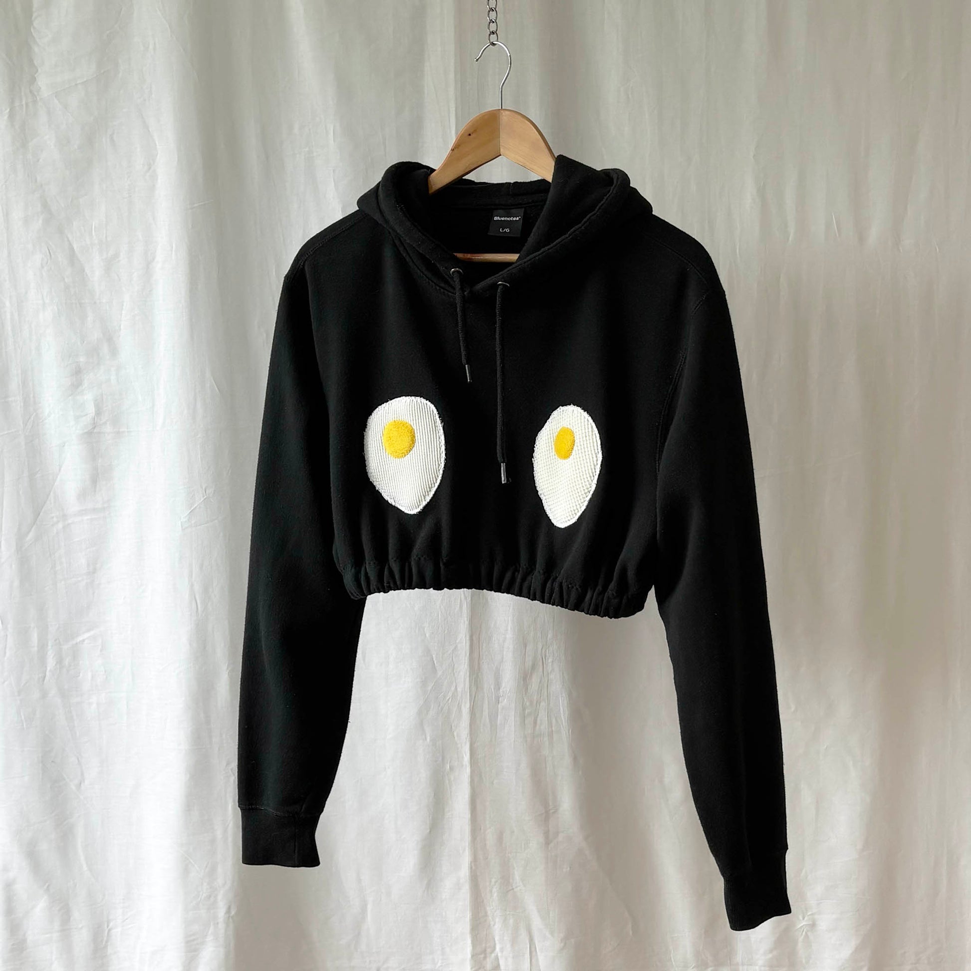 Cropped black hoodie with egg design on a hanger against a white background