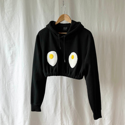 Cropped black hoodie with egg design on a hanger against a white background