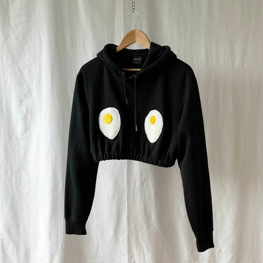 Cropped black hoodie with egg design on a hanger against a white background