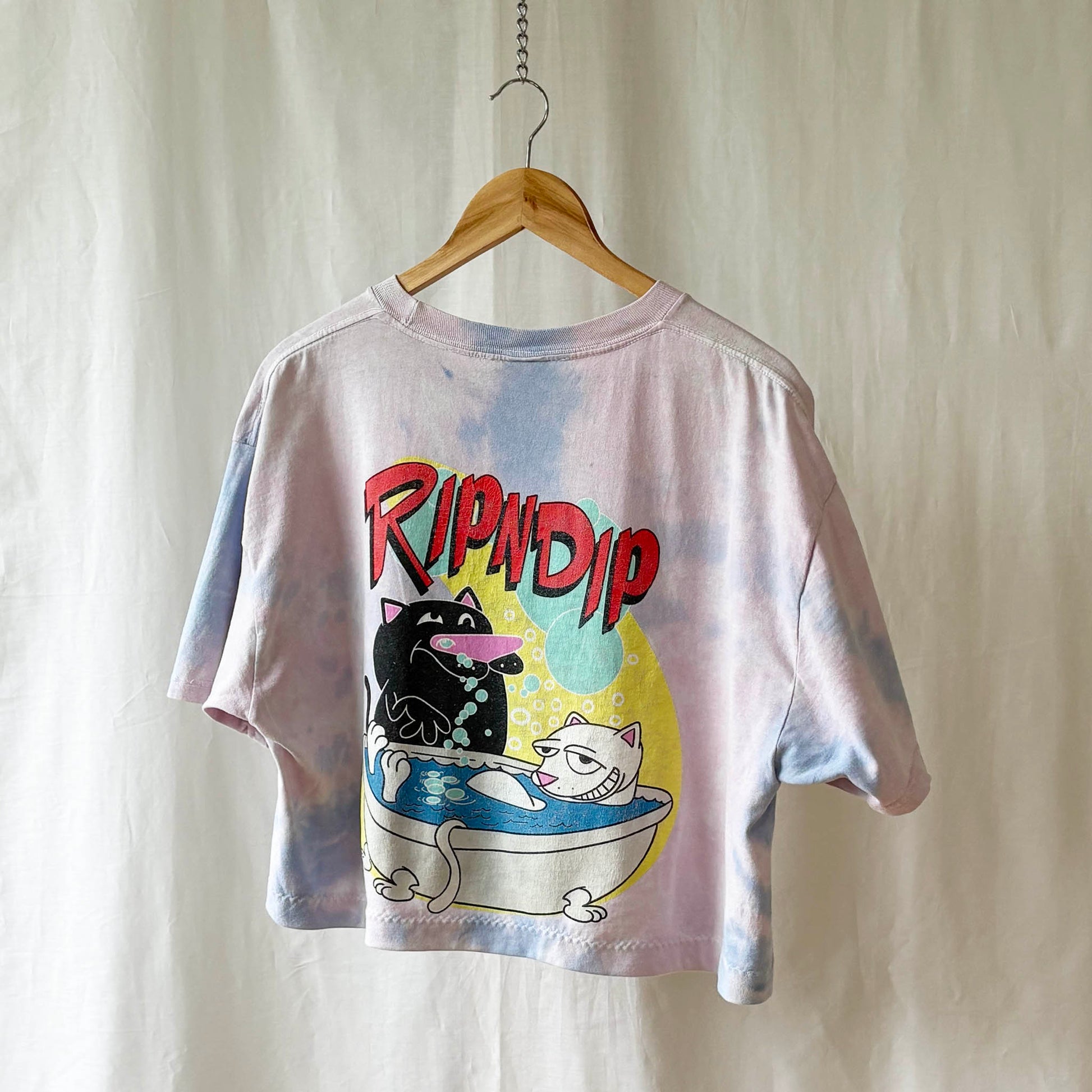 Tie-dye t-shirt with a cat farting in a tub an a brand logo on a wooden hanger against a plain background