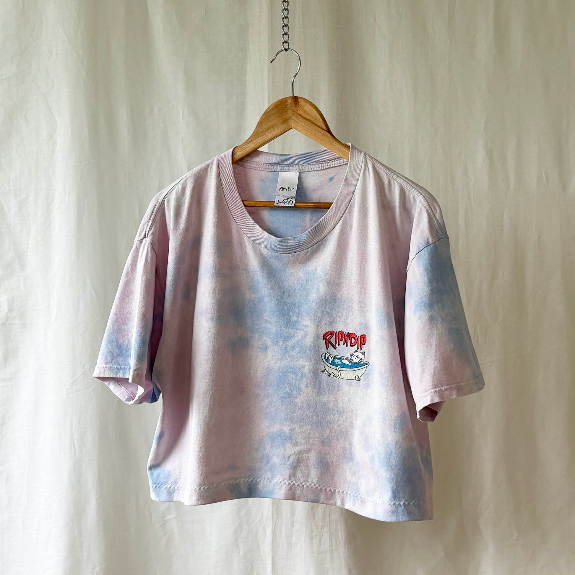 Tie-dye t-shirt with a cat farting in a tub an a brand logo on a wooden hanger against a plain background