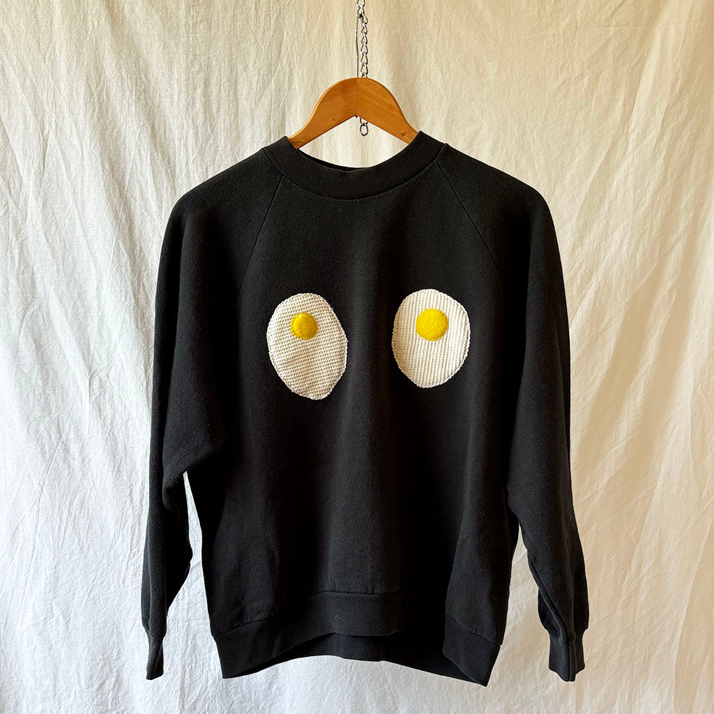 Black sweatshirt with egg design on a wooden hanger against a textured white background
