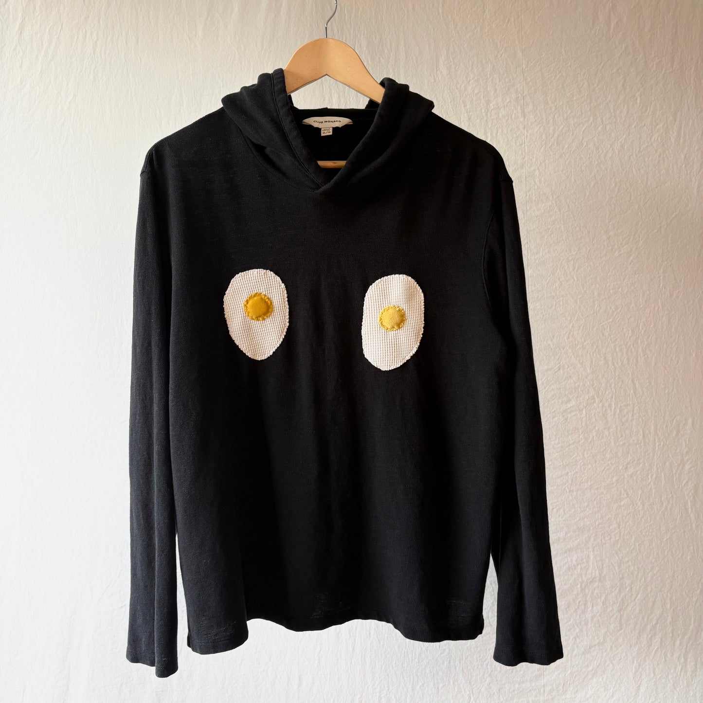A black knit hoodie hanging on a wooden hanger against a textured off-white wall. The sweater features two handmade egg patches on the chest, with white egg whites and yellow yolks.