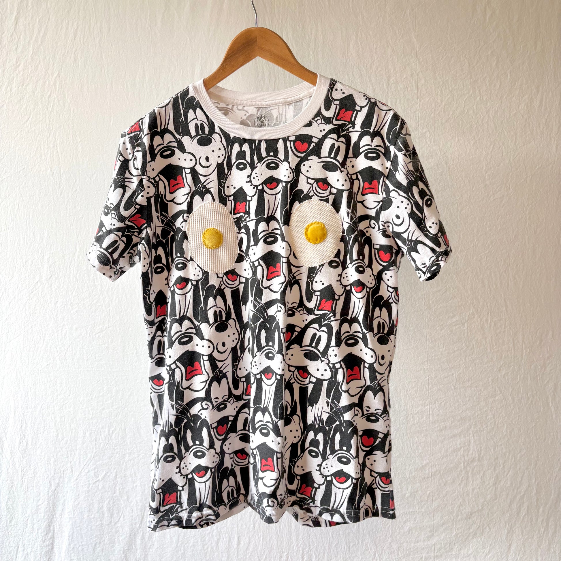 A short-sleeve crewneck t-shirt featuring an all-over black and white Goofy print with red accents, decorated with two 3D applique eggs on the chest, hanging on a wooden hanger against a textured off-white wall.