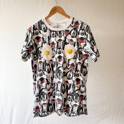 A short-sleeve crewneck t-shirt featuring an all-over black and white Goofy print with red accents, decorated with two 3D applique eggs on the chest, hanging on a wooden hanger against a textured off-white wall.