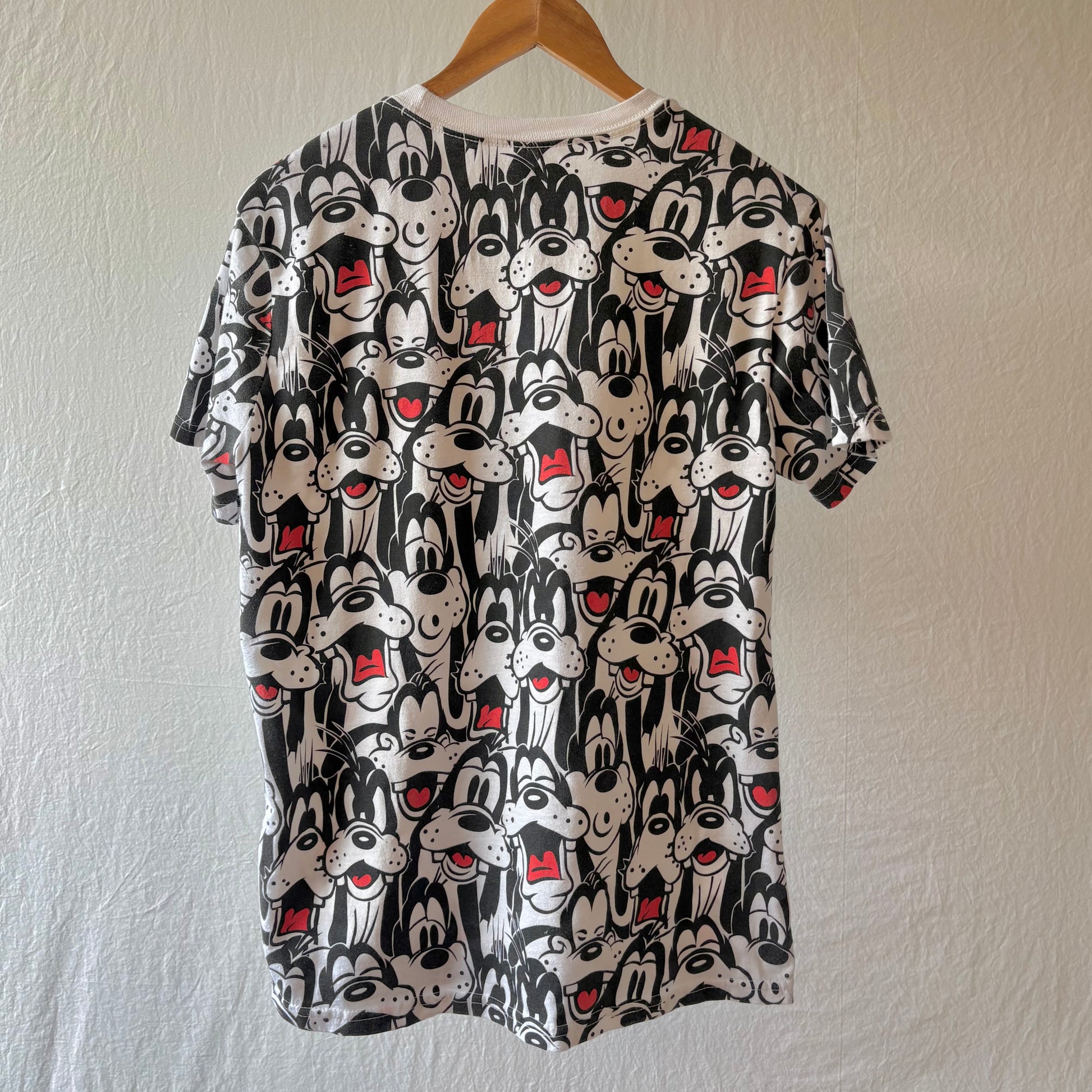 A short-sleeve crewneck t-shirt hanging on a wooden hanger against a textured off-white wall. The shirt features an all-over repeating print of Goofy faces in black and white with small red accents.