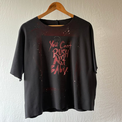 A black short-sleeve stenciled t-shirt hanging on a wooden hanger against a textured off-white wall. The shirt features a distressed red graphic on the front with the text 'You Can't Rush Art Baby.