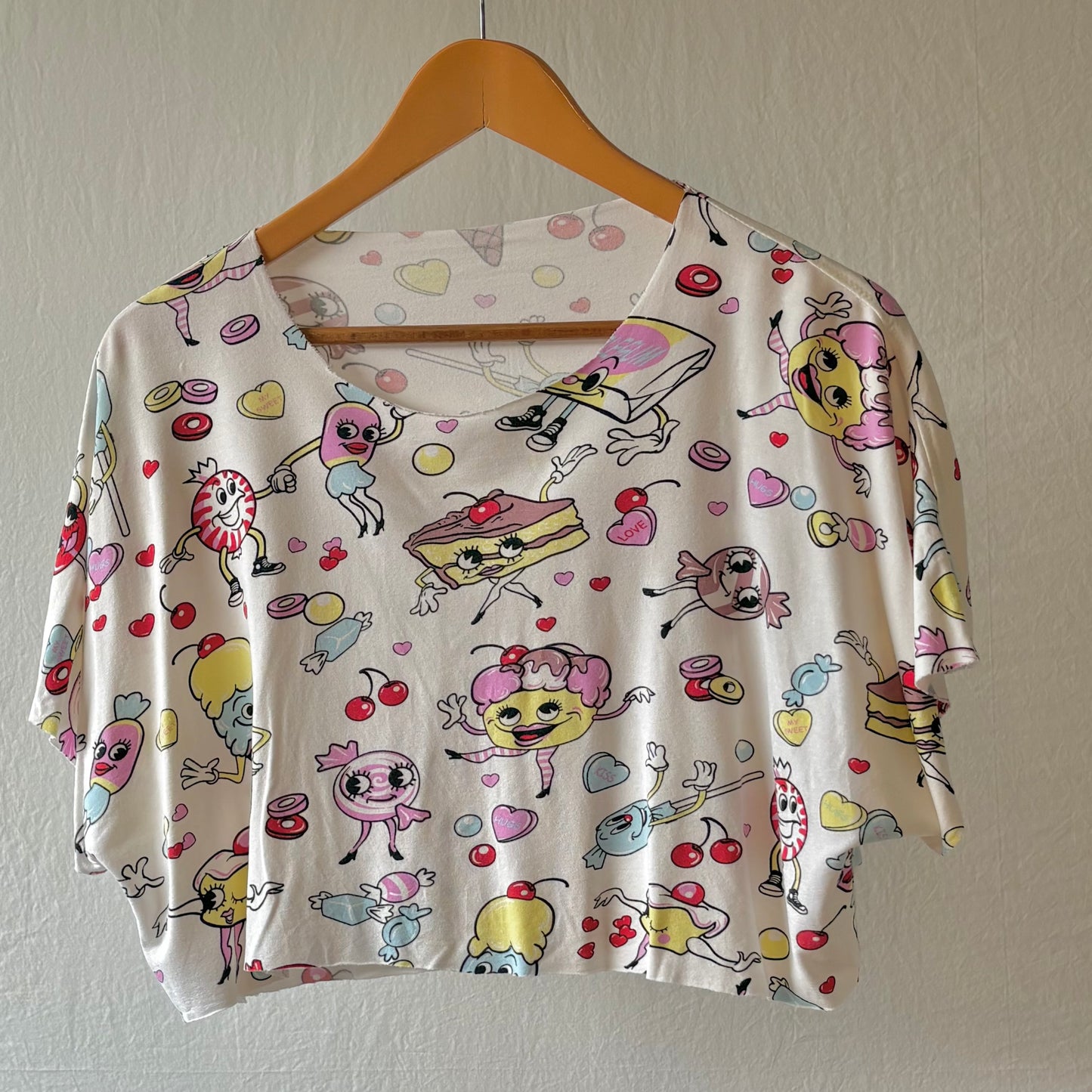 A short-sleeve crop top with a scoop neckline, featuring a playful cartoon print of characters, sweets, and hearts on an off-white background, hanging on a wooden hanger against a textured wall.