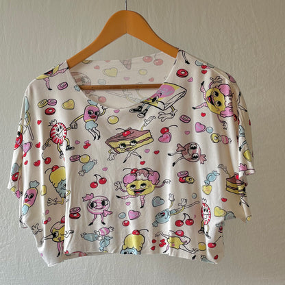A short-sleeve crop top with a scoop neckline, featuring a playful cartoon print of characters, sweets, and hearts on an off-white background, hanging on a wooden hanger against a textured wall.
