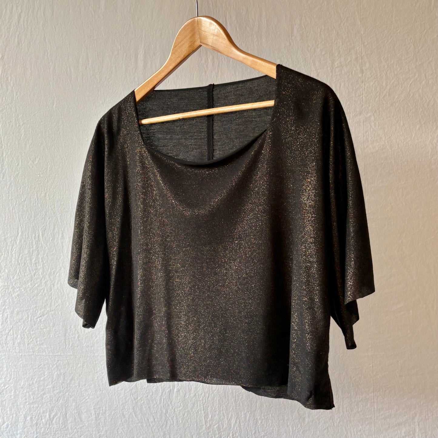 A black metallic shimmer short-sleeve top with a wide squared neckline, hanging on a wooden hanger against a textured off-white wall.