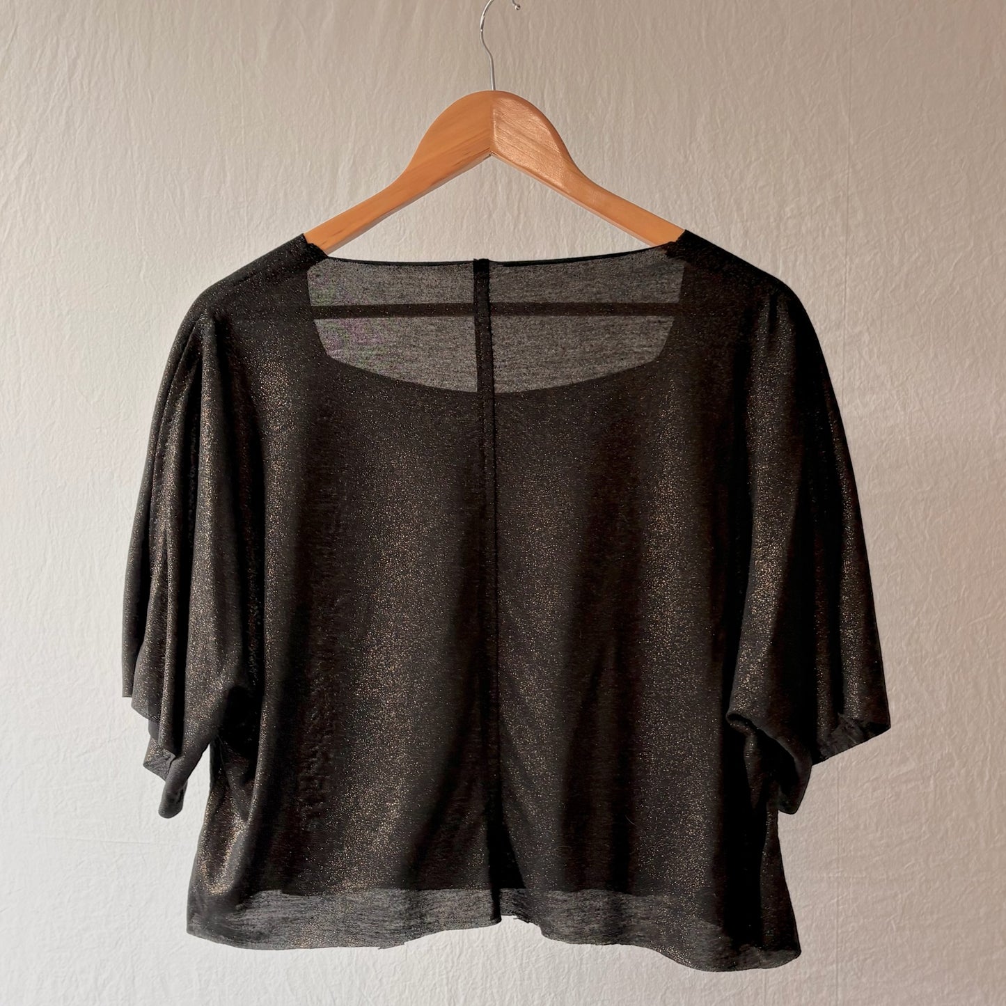 A black metallic shimmer short-sleeve top with a square neckline, hanging on a wooden hanger against a textured off-white wall.