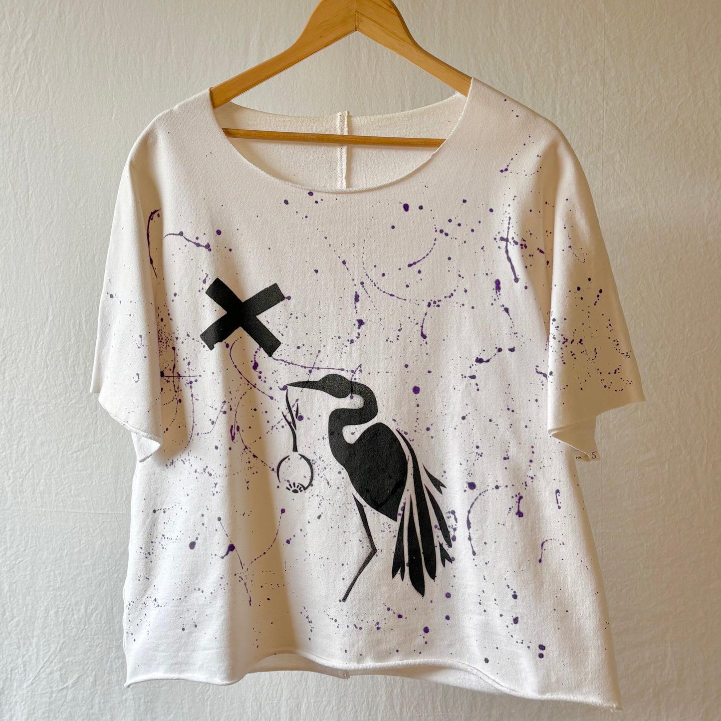 An off-white short-sleeve t-shirt with a black crane stencil and purple paint splatter details, hanging on a wooden hanger against a textured beige wall.