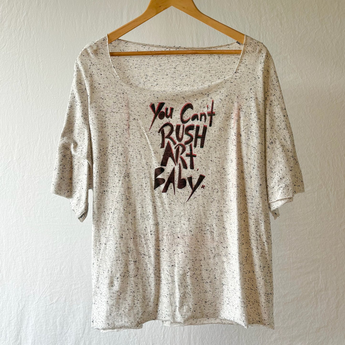 A short-sleeve scoop-neck t-shirt with a speckled cream and brown pattern, featuring the text 'You Can't RUSH ART BABY.' stenciled on the front, hanging on a wooden hanger against a textured white wall.