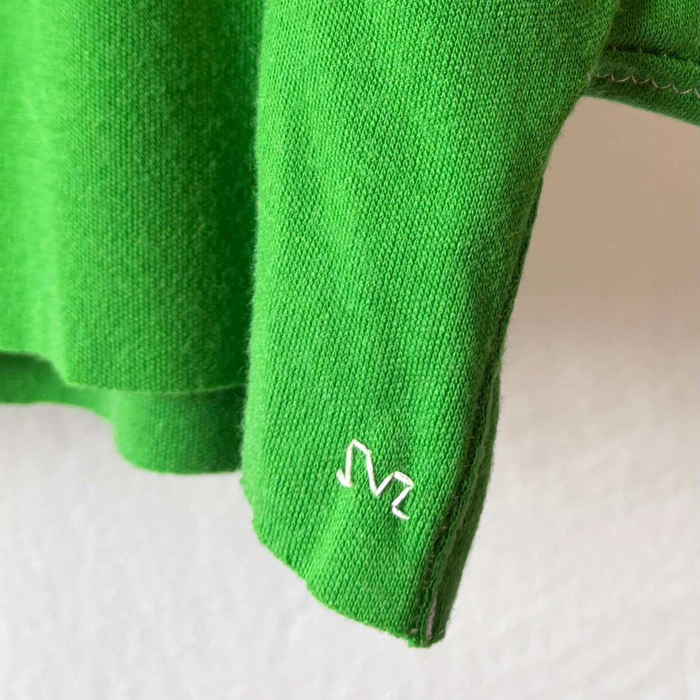 Close-up of a bright green knit garment, likely a sweater or scarf, showing a small white embroidered SVZ monogram logo on the bottom right raw edge against a plain light background.