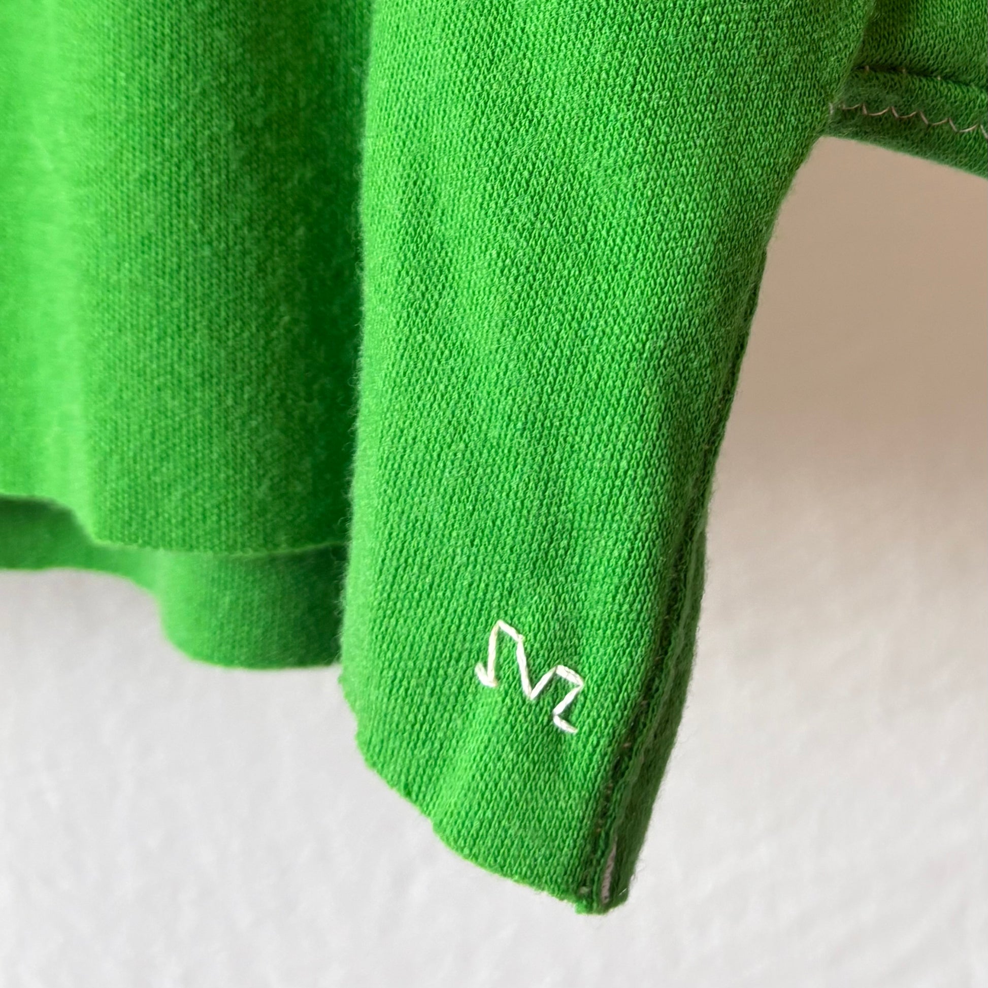 Close-up of a bright green knit garment, likely a sweater or scarf, showing a small white embroidered SVZ monogram logo on the bottom right raw edge against a plain light background.