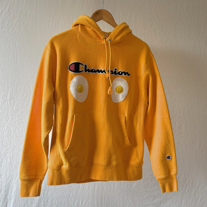 A bright yellow Champion hoodie hanging on a wooden hanger against a textured white wall. The hoodie features two handmade egg patches on the front with the Champion logo above it.