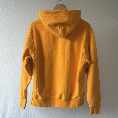 A bright yellow hoodie shown from the back, hanging on a wooden hanger against a textured off-white wall.