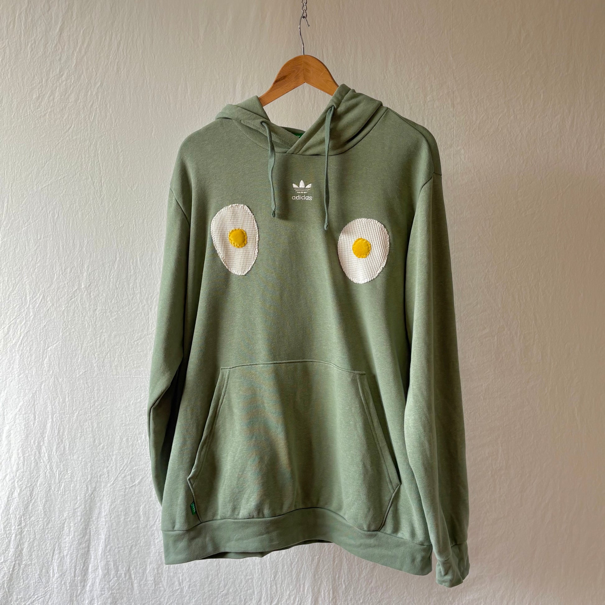 A sage green Adidas hoodie hanging on a wooden hanger against a textured off-white wall. The hoodie features a front kangaroo pocket and drawstrings, with two handmade egg patches on the chest.
