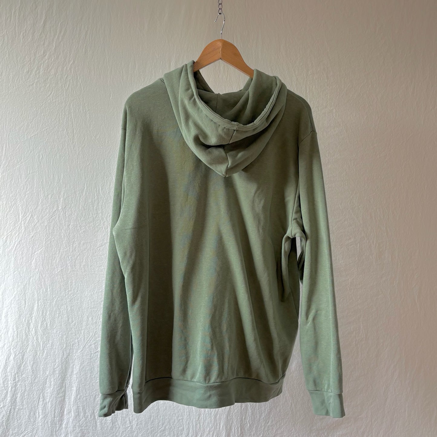 A sage green hooded sweatshirt hanging on a wooden hanger against a textured off-white wall.