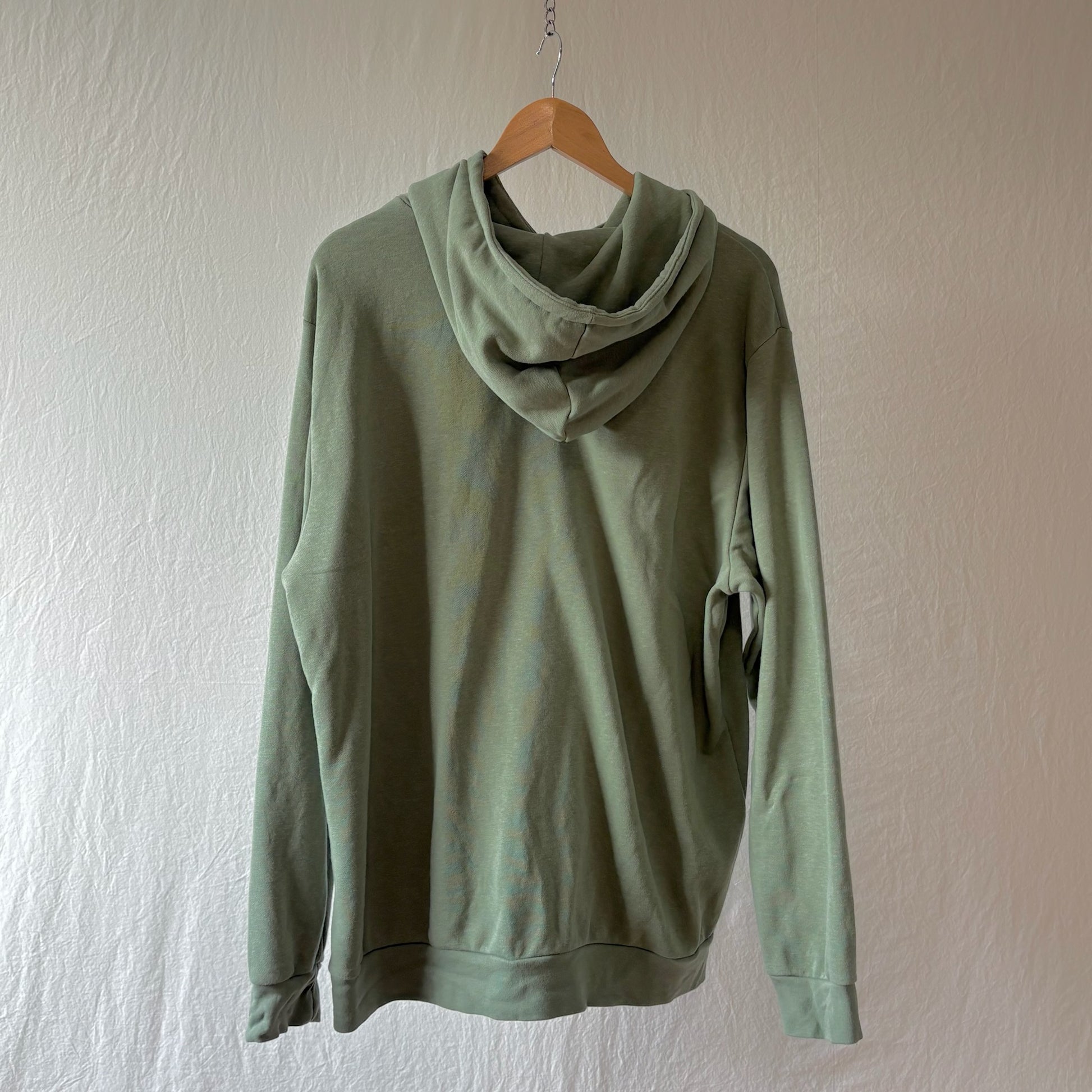 A sage green hooded sweatshirt hanging on a wooden hanger against a textured off-white wall.