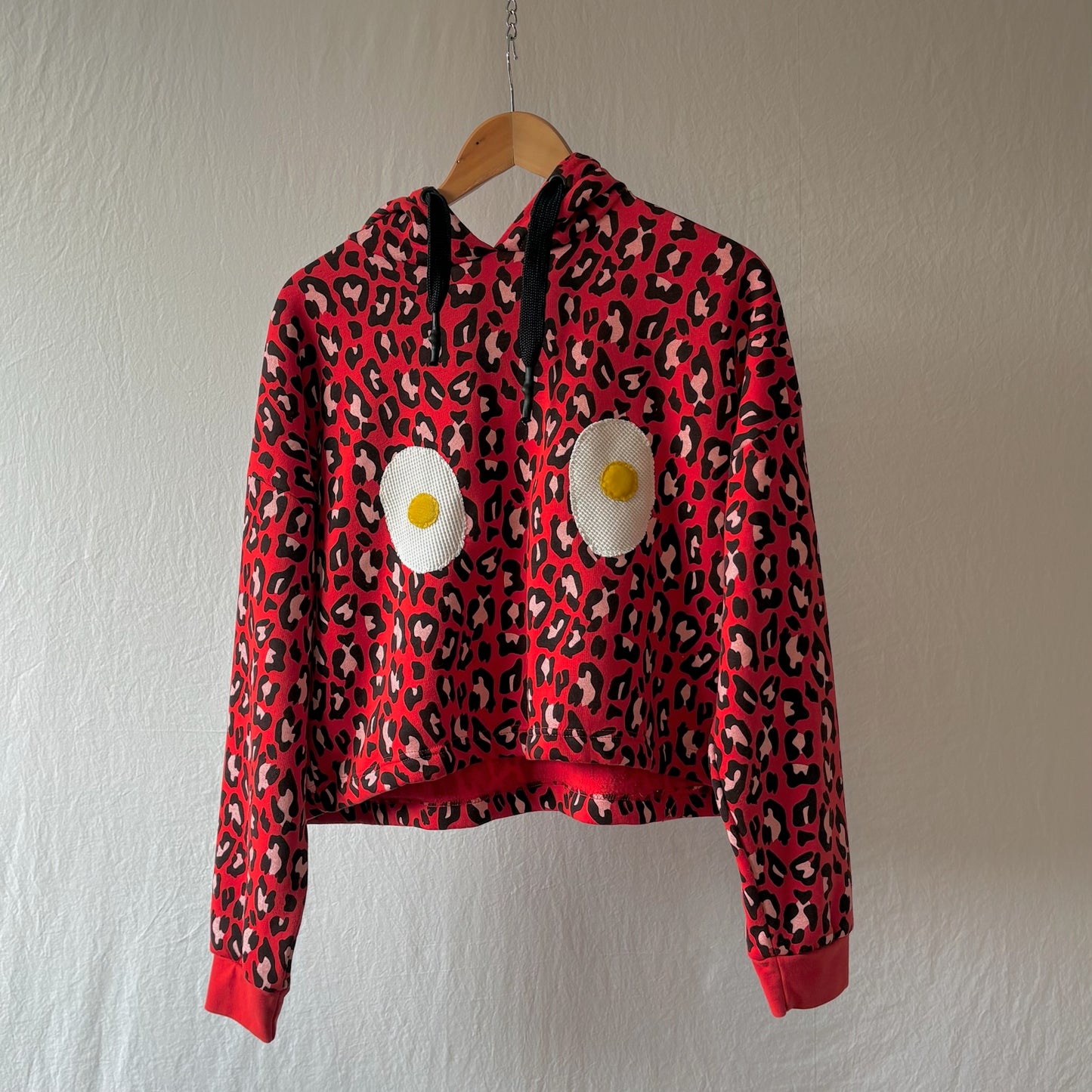 A red leopard-print cropped hoodie with black drawstrings and two large handmade egg patches on the front, hanging on a wooden hanger against a textured off-white wall.