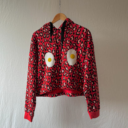 A red leopard-print cropped hoodie with black drawstrings and two large handmade egg patches on the front, hanging on a wooden hanger against a textured off-white wall.