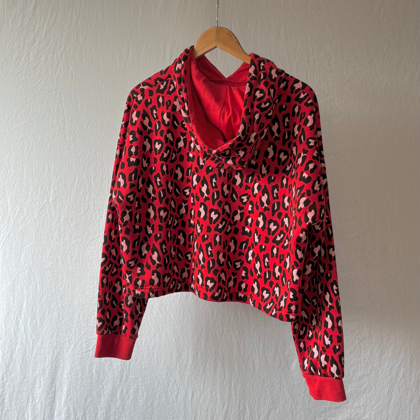 A red long-sleeve crop top hoodie with a black and beige leopard print pattern, hanging on a wooden hanger against a textured off-white wall.