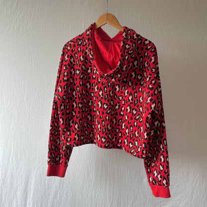 A red long-sleeve crop top hoodie with a black and beige leopard print pattern, hanging on a wooden hanger against a textured off-white wall.