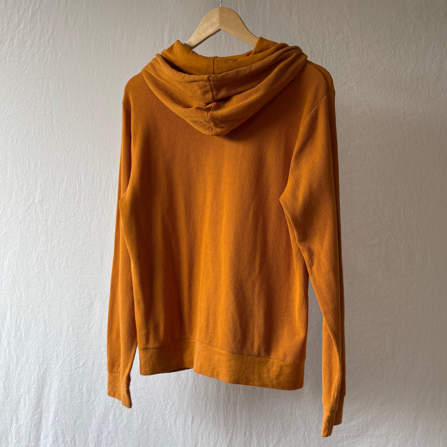 A golden yellow waffle weave hooded sweatshirt hanging on a wooden hanger against a textured off-white wall.