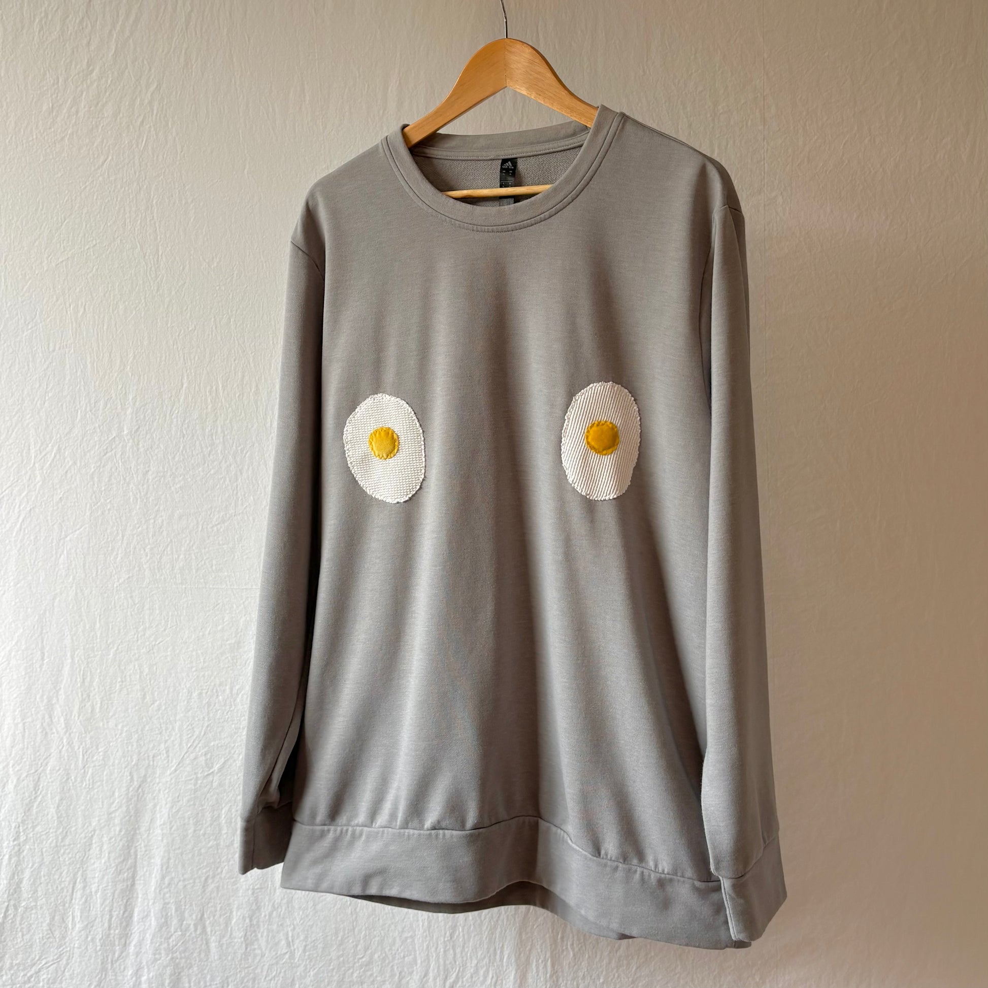 A light grey long-sleeve sweatshirt hanging on a wooden hanger against a textured off-white wall. The sweatshirt features two handmade egg patches on the chest.