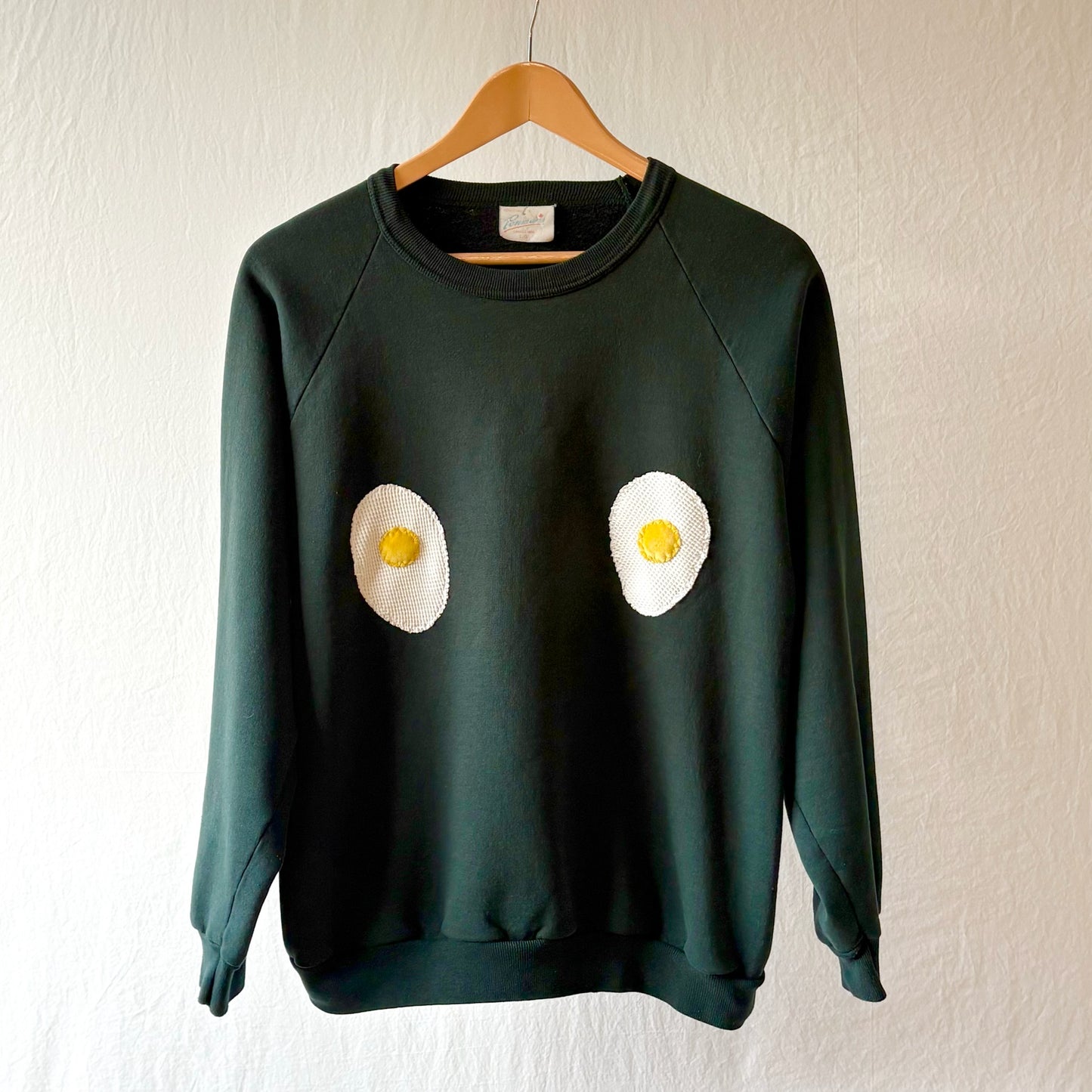 A dark green crewneck sweatshirt hanging on a wooden hanger against a textured off-white wall. The sweatshirt features two handmade egg patches on the chest, with white egg whites and yellow yolks.