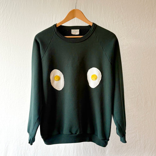 A dark green crewneck sweatshirt hanging on a wooden hanger against a textured off-white wall. The sweatshirt features two handmade egg patches on the chest, with white egg whites and yellow yolks.