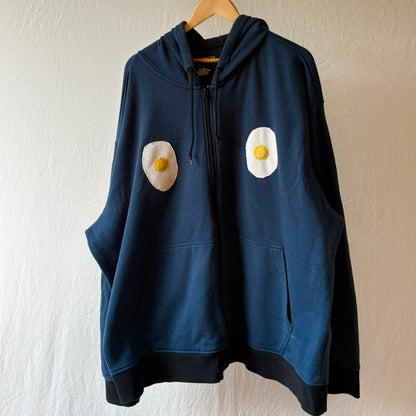 A navy blue zip-up hoodie hanging on a wooden hanger against a textured off-white wall. The hoodie features two hand made fried egg patches on the chest.