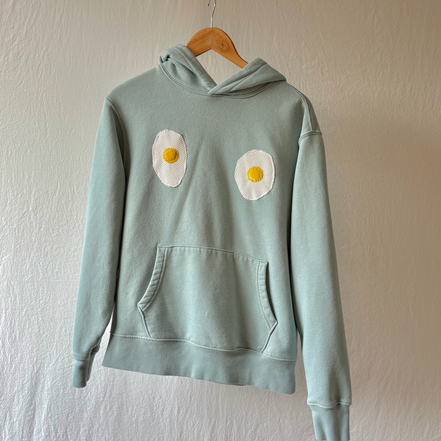 A slight teal pullover hoodie hanging on a wooden hanger against a textured off-white wall. The hoodie features two handmade fried egg patches on the chest and a front kangaroo pocket.