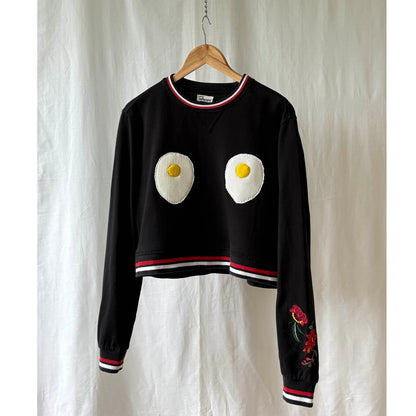 Black sweater with egg design  on chest and flowers embroidered on left sleeve on a hanger against a white background