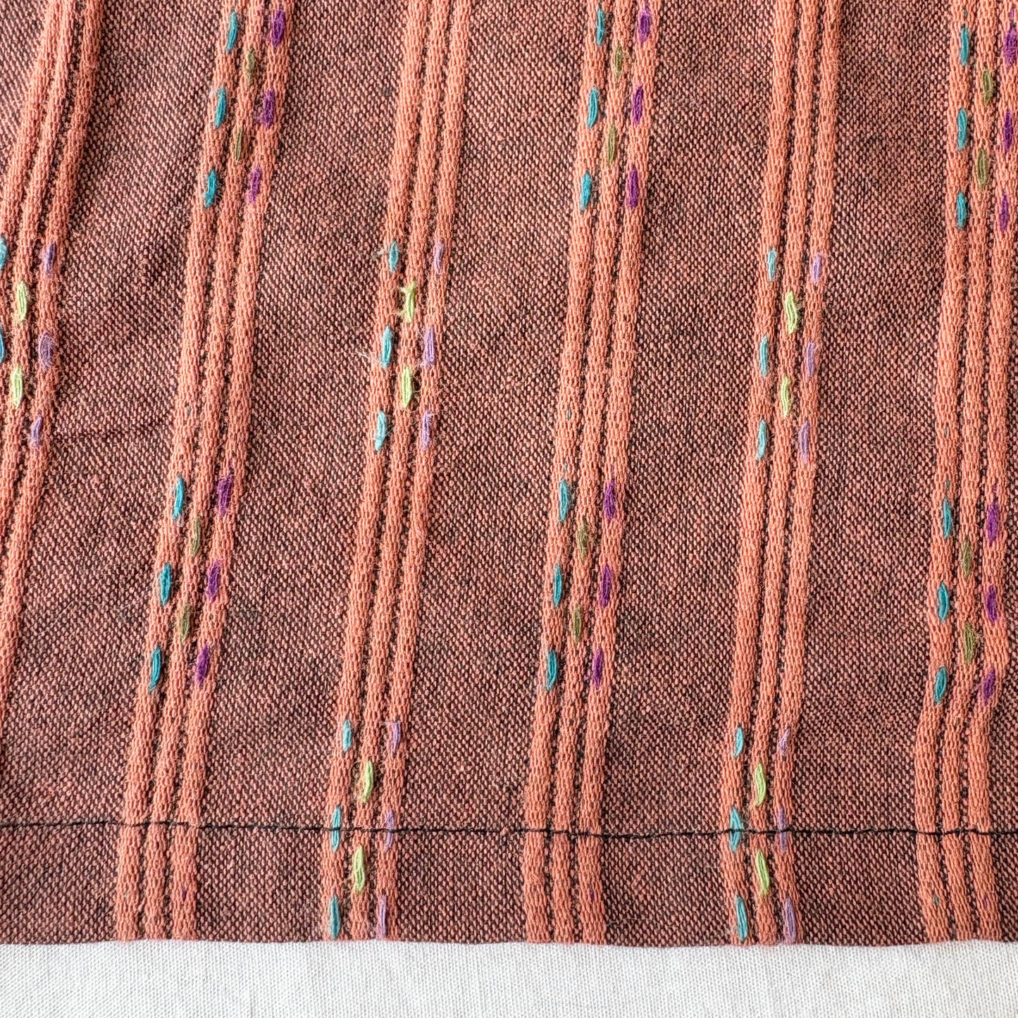 Close-up of orange fabric with teal, lime green and purple embroidered patterns on a white background